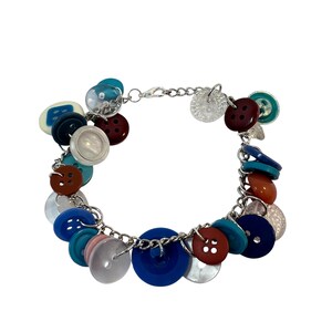 May include: A charm bracelet featuring a silver-toned metal chain adorned with an assortment of multi-colored plastic and glass buttons in shades of blue, red, brown, pink, and white, secured with a lobster claw clasp and an extender chain.