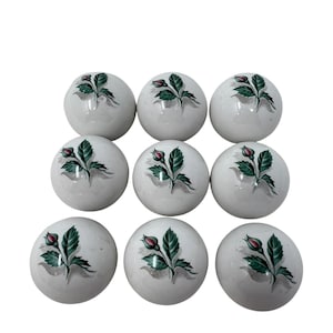 May include: Nine white ceramic cabinet knobs with a floral design. Each knob features a green leaf and red rose bud illustration. The knobs are round and have a glossy finish, perfect for adding a touch of elegance to furniture.
