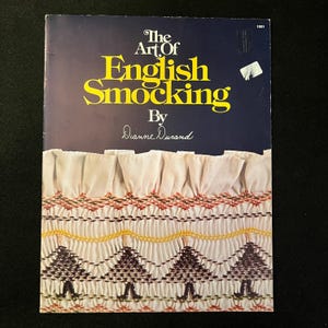 May include: A vintage book titled "The Art Of English Smocking" by Dianne Durand. The cover features a navy blue background with yellow text and a detailed smocked fabric design in white, brown, yellow, and green.