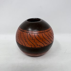 May include: A round, handcrafted pottery vase with a dark brown top and base, and a reddish-brown center section. The center features a repeating zigzag pattern. The vase has a small opening at the top and is set against a white background.