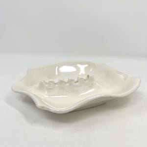 May include: Off-white ceramic ashtray with a wavy, irregular shape. The ashtray has a shallow bowl with several grooves for resting cigarettes. The glossy finish reflects light, highlighting the curves and contours of the design.