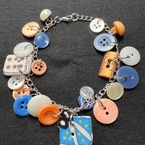 May include: A handmade charm bracelet featuring a variety of colorful buttons in shades of blue, orange, white, and pink. The bracelet includes a charm with scissors and a blue fabric design. The bracelet is made with a silver chain.