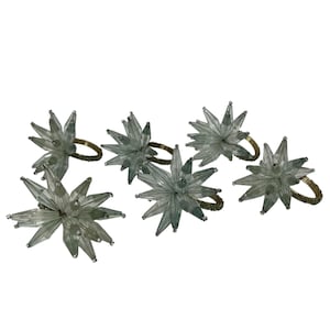 May include: Six star-shaped napkin rings. Each ring features a clear, spiky star design with a gold-colored ring base. The napkin rings are arranged on a white background, showcasing their decorative appeal for table settings.