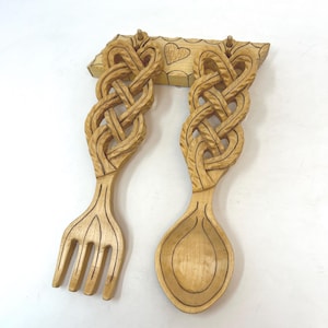 May include: Wooden fork and spoon set with intricate Celtic knotwork design. The light-colored wood utensils are carved with detailed patterns and feature a heart-shaped accent on the connecting piece. A decorative kitchen item.
