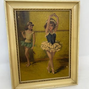 May include: Vintage framed print of two young ballerinas in a dance studio. The artwork features a ballerina in a blue and cream tutu with arms raised, and another in a green tutu holding a flower. The frame is a light beige color with a gold trim.
