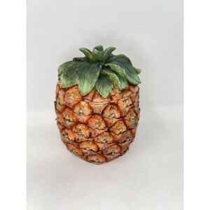 May include: A decorative pineapple-shaped container. The container is a warm orange-brown color with a textured surface, mimicking the skin of a pineapple. The top features green leaves, adding a naturalistic touch. This item is ideal for kitchen decor.