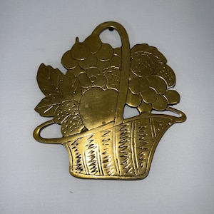 May include: A brass-colored metal basket overflowing with fruit. The basket features grapes, strawberries, and an apple. The handle curves upward, and the basket has a textured, woven appearance. A decorative piece.