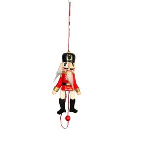 May include: A wooden nutcracker ornament in the shape of a soldier. The ornament has a red jacket, black boots, and a black hat with a gold accent. It has a white beard and mustache. The ornament hangs from a red string.