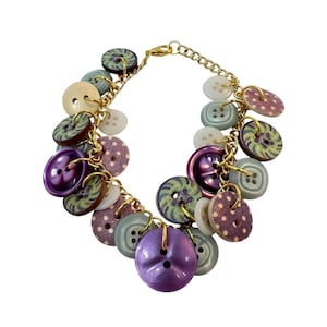 May include: A charm bracelet featuring a gold-tone metal chain adorned with an assortment of multi-colored buttons. The buttons vary in size, color, and pattern, including solid purple, light blue, white, and patterned designs with dots and swirls.