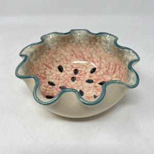 May include: A ceramic bowl with a watermelon design. The interior features a pink and white speckled pattern with black seed details. The rim is scalloped with a blue trim, and the exterior is off-white.