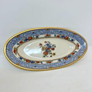 May include: Oval-shaped ceramic serving dish with a gold rim and floral design. The dish features a light blue border with a repeating pattern and a central floral arrangement in red, blue, and yellow.