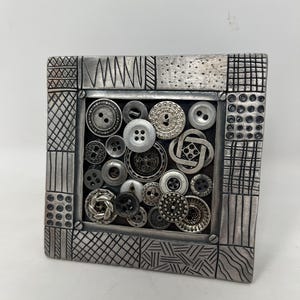 May include: A square, silver-toned frame filled with an assortment of buttons in various sizes, shapes, and textures. The frame has a textured border with geometric patterns. The buttons are in shades of silver and gray.