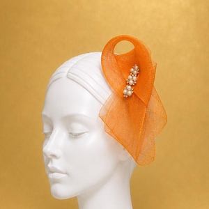 May include: An orange fascinator hat made of sheer, stiffened fabric shaped into a looped, sculptural design. The accessory is embellished with a vertical row of small, round, white faux pearls set against the vibrant orange mesh material.