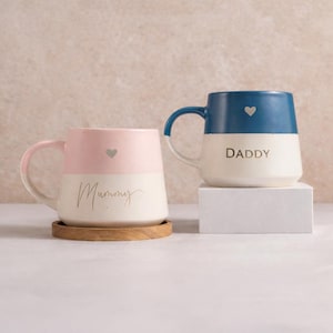 Puede incluir: Dos tazas de cerámica con diseño bicolor. La taza de la izquierda es rosa claro y crema con la palabra Mummy en letra dorada y un pequeño corazón. La taza de la derecha es azul marino y crema con la palabra DADDY en letras doradas y un corazón.