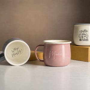 Pode incluir: Três canecas de estilo esmaltado: uma caneca cinzenta deitada de lado com o texto "Will you Marry Me?" no interior, uma caneca rosa com texto branco em cursivo e uma caneca creme com rebordo cinzento e o texto "Say YES to new ADVENTURES".