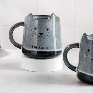 Könnte beinhalten: Zwei Keramikbecher in Katzenform. Der Becher links ist grau mit schwarzen Streifen und trägt die Aufschrift "Cat Mum". Der Becher rechts ist grau mit schwarzen Streifen und hat ein schwarzes Katzen Gesicht.