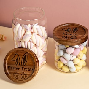 May include: Three glass jars with wooden lids. The lids are engraved with bunny ears and the words "Easter Treats", "Kieran", and "Samuel". The jars are filled with colorful candies and marshmallows.