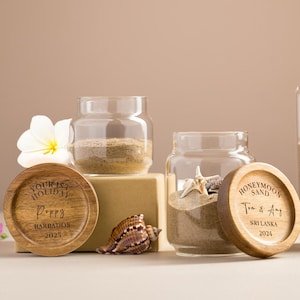 Pode incluir: Três frascos de vidro cheios de areia e conchas, cada um com uma tampa de madeira. As tampas são gravadas com texto, incluindo "Honeymoon Sand" e "Tourist Holiday". Uma flor branca e uma flor rosa também estão presentes.