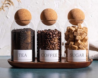 Puede incluir: Tres tarros de vidrio con tapas de corcho y etiquetas que dicen "TÉ", "CAFÉ" y "AZÚCAR". Los tarros están llenos de hojas de té, granos de café y cubos de azúcar, respectivamente. Los tarros están sobre una bandeja de madera.