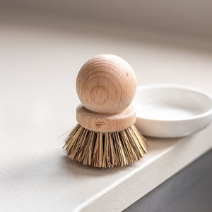 Może przedstawiać: Drewniana szczotka do mycia naczyń z naturalnym włosiem, leżąca na białym blacie obok białej ceramicznej miski.