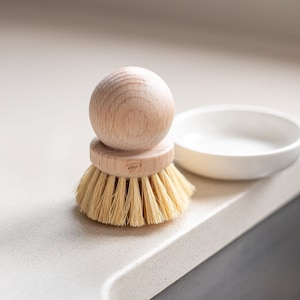 Könnte beinhalten: Eine Holzbürste mit rundem Griff und Naturborsten, neben einer kleinen weißen Keramikschale. Die Bürste ist hellbraun und beige, und die Schale ist rund. Die Gegenstände befinden sich auf einer hellen Arbeitsplatte.