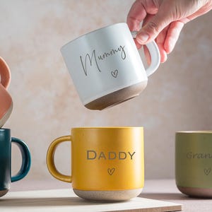 May include: A selection of ceramic mugs in shades of orange, teal, yellow, white, and green. Each mug has a contrasting base and features a word and a heart symbol. The words on the mugs are "Coffee", "Pop", "Daddy", "Grandma", and "Mummy".