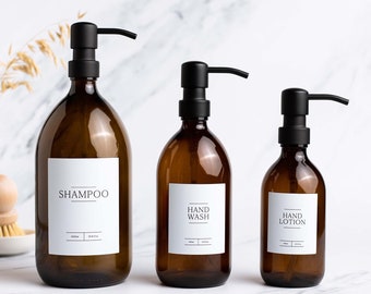 Può includere: Tre bottiglie di vetro ambrato con pompe nere e etichette bianche. Le etichette riportano "Shampoo", "Lavamani" e "Lozione per le mani".