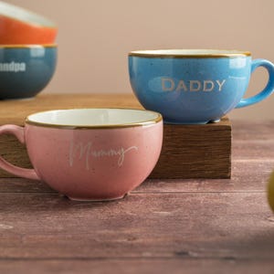 May include: A selection of ceramic mugs in shades of pink, blue, and orange. The mugs are speckled and inscribed with the words "Daddy," "Mummy," and "Grandpa." Each mug has a handle and a gold trim.