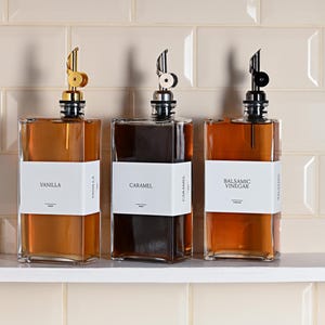 May include: Three glass bottles with gold, white, and black pour spouts, filled with liquids. The bottles are labeled "Vanilla", "Caramel", and "Balsamic Vinegar".