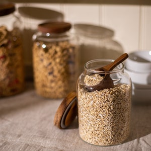 May include: Three glass jars with wooden lids filled with different types of cereal. The jar in the foreground is filled with rolled oats and has a wooden spoon inside.