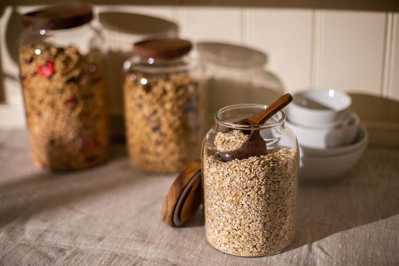 May include: Three glass jars with wooden lids filled with different types of cereal. The jar in the foreground is filled with rolled oats and has a wooden spoon inside.