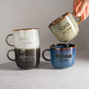 Puede incluir: Cuatro tazas de cerámica con asas y bordes marrones oscuros. Las tazas son de varios colores: blanco, gris oscuro, verde moteado y azul. Cada taza tiene un nombre y el año 2026: Mummy, Daddy, Nana y Grandad.