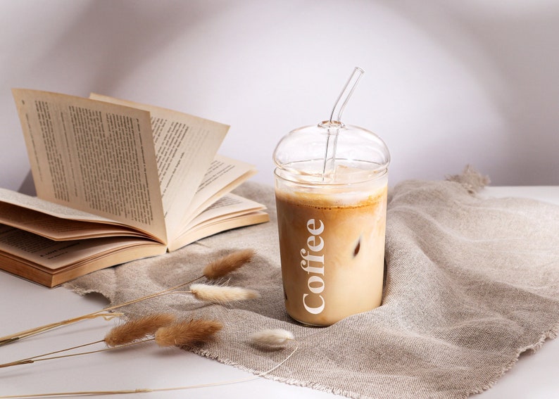 Pu&ograve; includere: Un bicchiere in vetro trasparente con coperchio a cupola e cannuccia in vetro abbinata, riempito di caff&egrave; freddo. La parola Coffee &egrave; stampata in lettere bianche verticali sul lato del bicchiere. Il bicchiere &egrave; posizionato accanto a un libro.