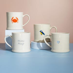 Może przedstawiać: Cztery kremowe ceramiczne kubki z różnymi wzorami. Jeden kubek ma kraba, drugi ptaka, trzeci ma wycięte serce, a ostatni napis "Mini Mug". Kubki stoją na niebieskiej powierzchni.
