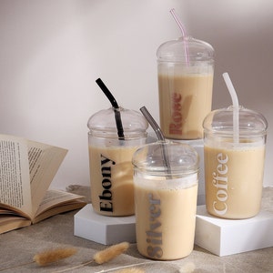 Pu&ograve; includere: Quattro bicchieri in vetro trasparente con coperchi a cupola e cannucce abbinate, ciascuno riempito con un liquido beige. I bicchieri presentano etichette con testo stampato che riportano Ebony, Silver, Rose e Coffee in colori distinti, tra cui nero, argento, rosa e bianco.