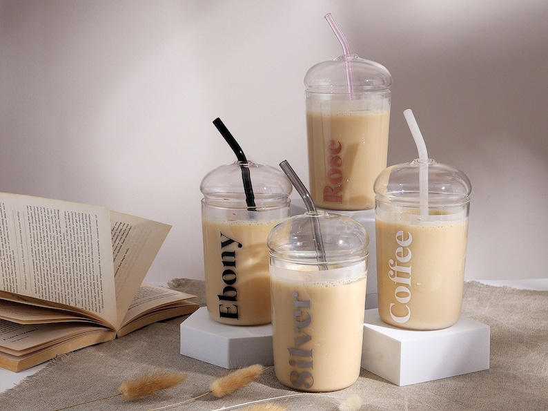 Pu&ograve; includere: Quattro bicchieri in vetro trasparente con coperchi a cupola e cannucce abbinate, ciascuno riempito con un liquido beige. I bicchieri presentano etichette con testo stampato che riportano Ebony, Silver, Rose e Coffee in colori distinti, tra cui nero, argento, rosa e bianco.