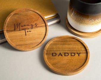 May include: Wooden coasters with engraved text. One coaster reads "Mummy's Tea" and another reads "DADDY". A mug with a brown and white gradient sits on a matching coaster. The coasters are made of light brown wood.