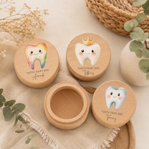 Può includere: Tre scatole rotonde in legno per dentini con coperchi. Ogni coperchio presenta un'illustrazione di un dente a cartone animato e il testo "TOOTH FAIRY BOX" con un nome sotto. Un dente è color arcobaleno, uno indossa una corona d'oro, uno è blu.