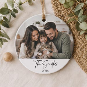 Op de afbeelding: Een rond wit keramieken ornament met een afgedrukte foto van een gezin van drie dat een kat vasthoudt. Onder de foto staat de tekst The Smiths en het jaartal 2026 in zwarte inkt. Het ornament hangt aan een lus van bruin jute touw.