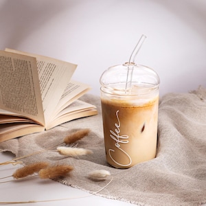 Pu&ograve; includere: Un bicchiere di vetro trasparente con coperchio a cupola e cannuccia di vetro abbinata, riempito con un caff&egrave; ghiacciato. La parola Coffee &egrave; scritta in corsivo bianco sul lato del bicchiere, posizionato accanto a un libro aperto.