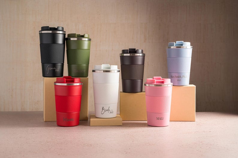 Op de afbeelding: Een verzameling ge&iuml;soleerde reismokken in verschillende kleuren, waaronder zwart, groen, rood, wit, bruin, roze en lavendel. Elke mok heeft een geribbelde siliconen hoes en een roestvrijstalen deksel. Sommige mokken hebben gepersonaliseerde tekst.