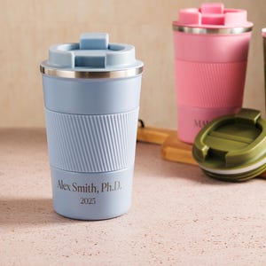 Op de afbeelding: Een verzameling ge&iuml;soleerde reismokken in verschillende kleuren, waaronder blauw, roze, groen en bruin. De blauwe mok heeft de tekst "Alex Smith, Ph.D. 2025". Elke mok heeft een geribbelde siliconen grip en een bijpassende deksel.