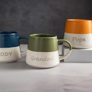 May include: Three ceramic mugs with a two-tone design featuring a speckled cream base and solid coloured tops in navy blue, olive green, and orange. Each mug is engraved with the text DADDY, Grandad, or Pops in a simple, clean font style.