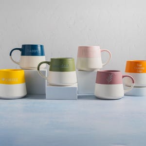 May include: A collection of six ceramic mugs, each featuring a two-tone design with a white base and a colored upper section. The mugs are in yellow, dark blue, olive green, light pink, mauve, and orange, with various text and icons printed on them.