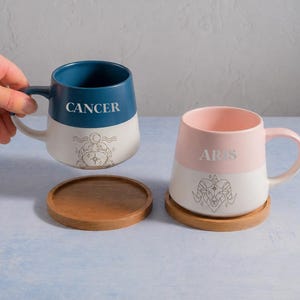 May include: Two ceramic mugs with matching wooden coasters. The left mug is dark blue and white with the text CANCER and a crab zodiac graphic. The right mug is light pink and white with the text ARIES and a ram zodiac graphic. Both have handles.