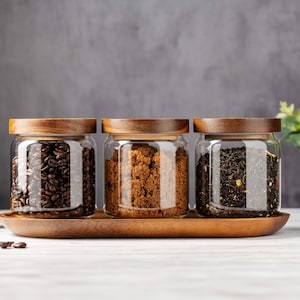 以下が含まれることがあります： コーヒー豆、ブラウンシュガー、茶葉が入った木製の蓋が付いたガラス瓶が3つ。瓶は木製のトレイの上に置かれています。