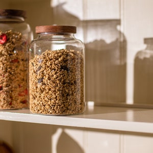 May include: Two glass jars with wooden lids filled with granola. The jar on the left contains red dried fruit in the granola. The jars are on a white shelf.