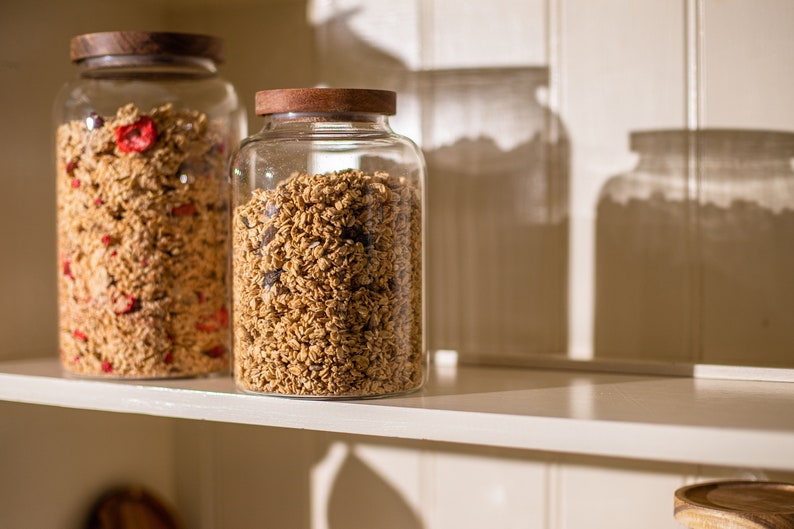 May include: Two glass jars with wooden lids filled with granola. The jar on the left contains red dried fruit in the granola. The jars are on a white shelf.
