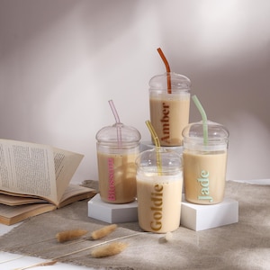 Pu&ograve; includere: Quattro bicchieri in vetro trasparente con coperchi a cupola e cannucce in vetro abbinate, riempiti con una bevanda beige. I bicchieri presentano i nomi Blossom, Amber, Goldie e Jade stampati nei colori corrispondenti sulla parte anteriore.
