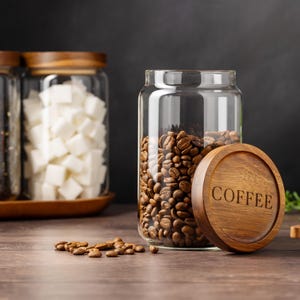 以下が含まれることがあります： コーヒーと書いてある木製の蓋が付いたガラスの瓶にコーヒー豆が入っています。背景には、木製の蓋が付いたガラスの瓶がもう2つあります。1つの瓶には砂糖の角砂糖が、もう1つの瓶には紅茶が入っています。